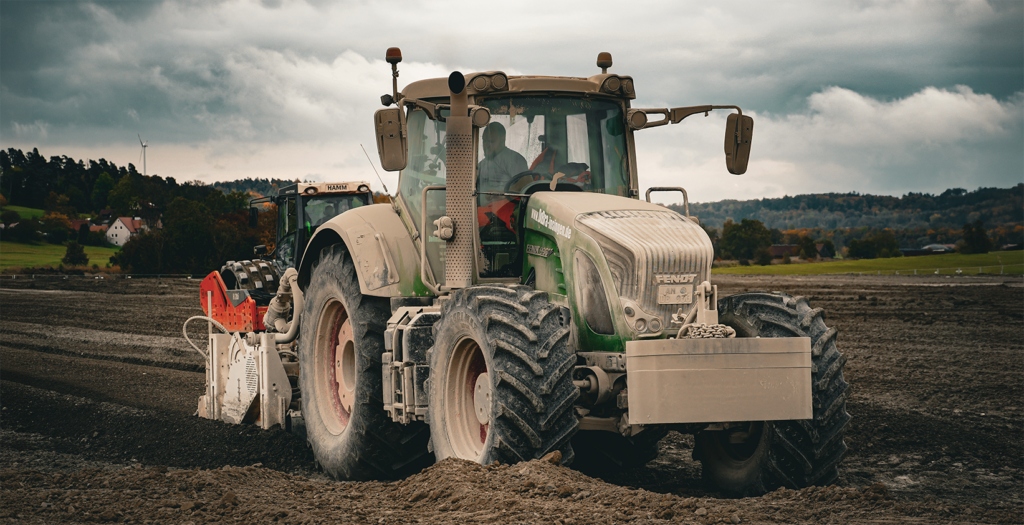 Fendt-Traktor mit Anbaugerät steht auf einem Erdfeld, im Hintergrund eine Windkraftanlage